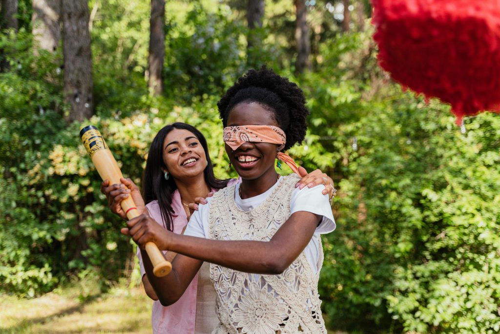 Two women laughing and playing a blindfolded game with a bat outdoors on a sunny day.